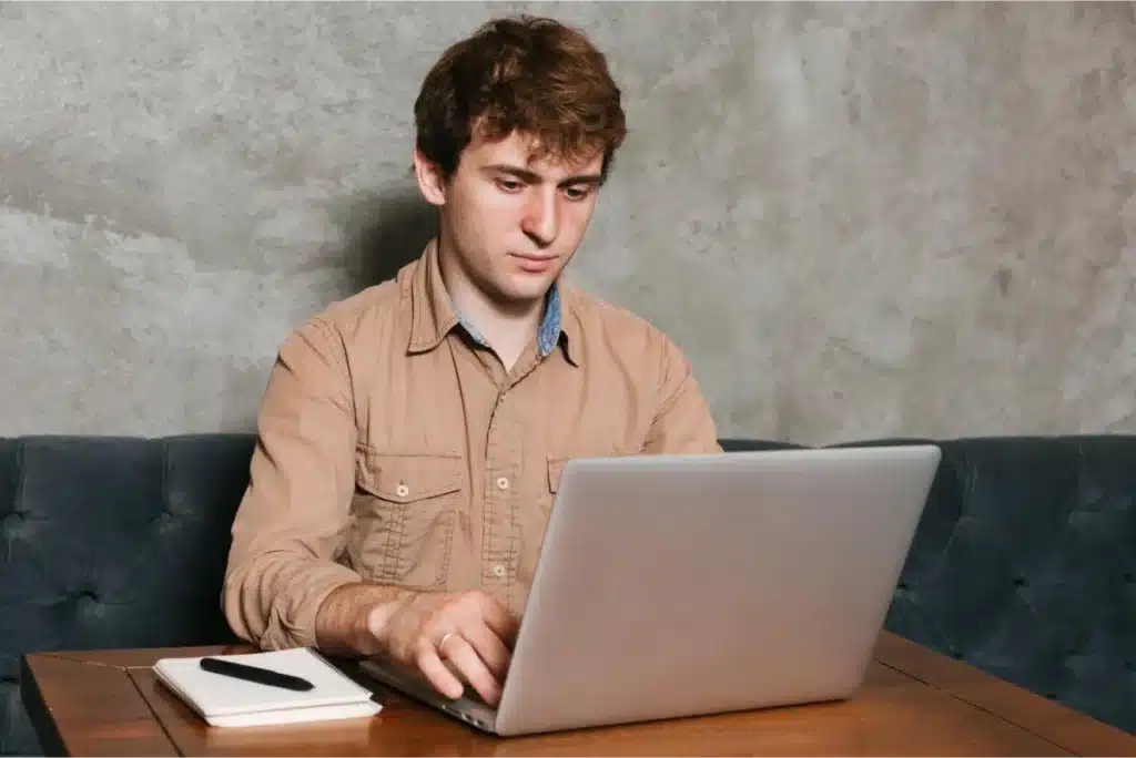 Man in a tan shirt working on a laptop at a wooden table, concentrating on his task with a notepad and pen nearby.