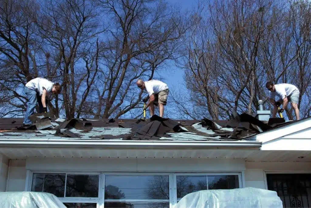 Three men removing old shingles from a roof, working together under a clear blue sky with bare trees in the background.