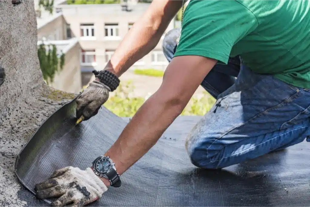 Construction worker in a green shirt applying a protective membrane on a roof, securing it around the base of a chimney.